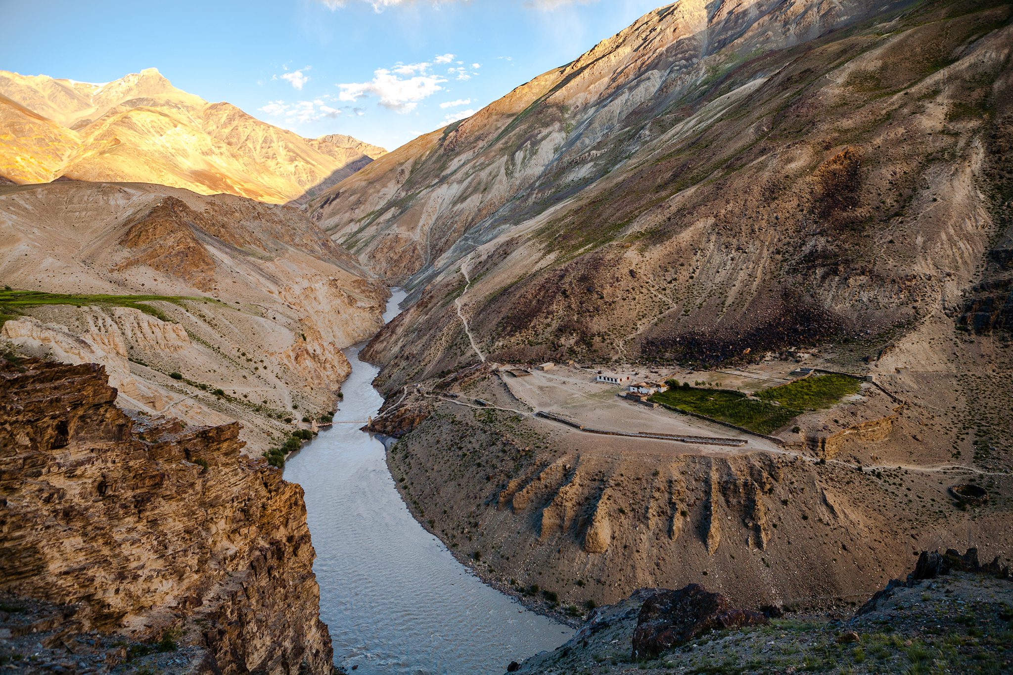 Cha village seen at a distance surrounded by the green farms with a rickety wooden bridge over river Lungnak to cross over to Purne is a homestay option for the first night halt on the trek to the remote and isolated Phuktal or Phugtal monastery or gompa passing through a deep gorge or ravine formed by the Lungnak, a tributary of the Indus river between the brown, golden and barren Greater Himalayan and Zanskar mountain ranges in the offbeat Zanskar valley in Jammu and Kashmir in North India.